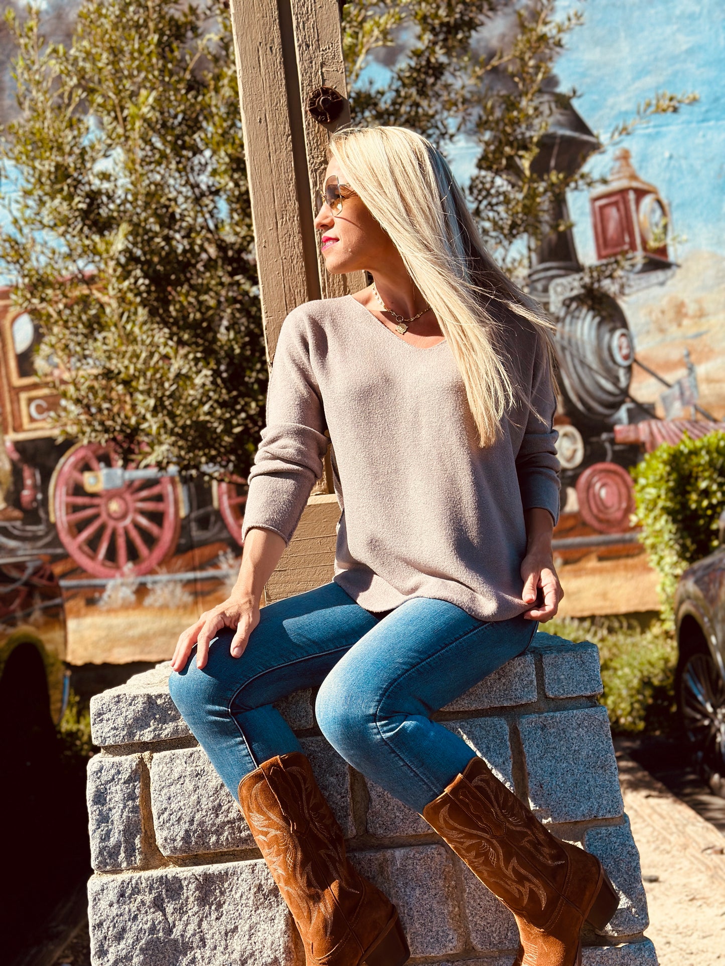 Woman sitting on stone steps wearing a beige sweater and blue jeans with brown boots, with a colorful background.