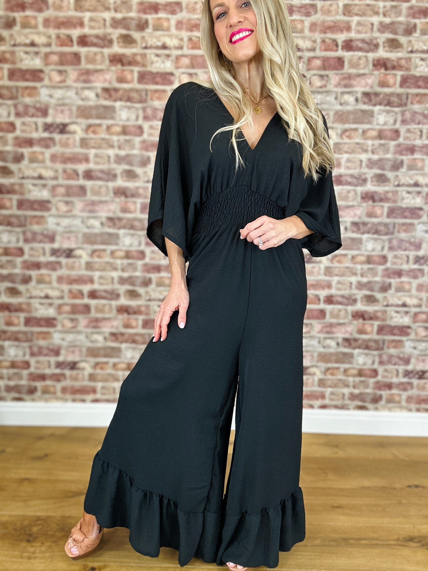A woman wearing a black jumpsuit with a deep V-neckline, loose fit, and frill detail at the ankle.
