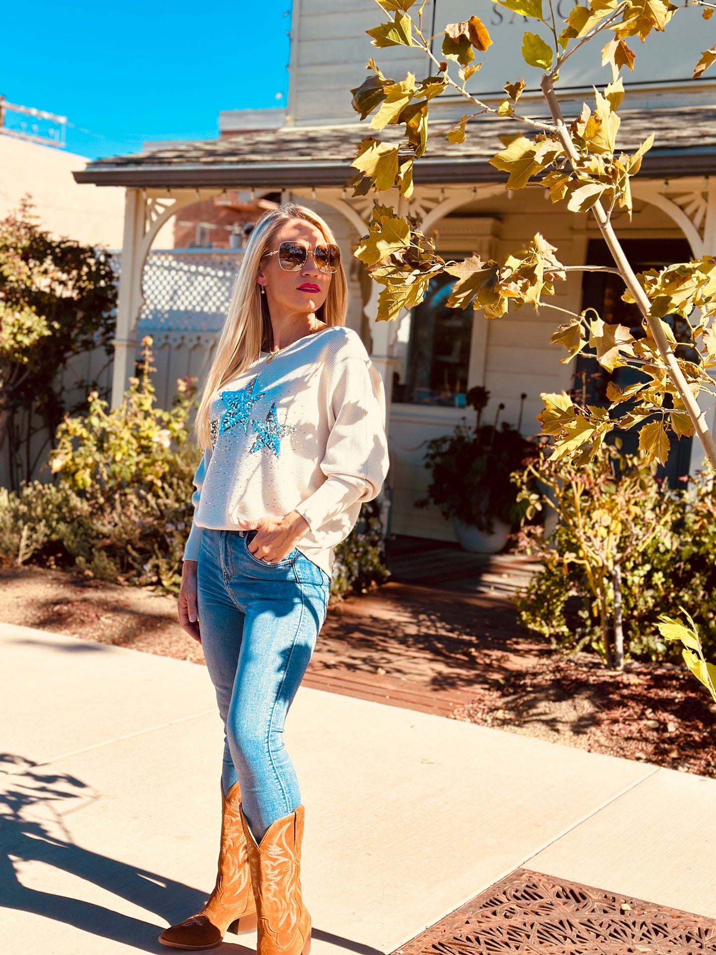 Woman in white sweater with blue floral design, blue jeans, and brown boots standing outdoors.