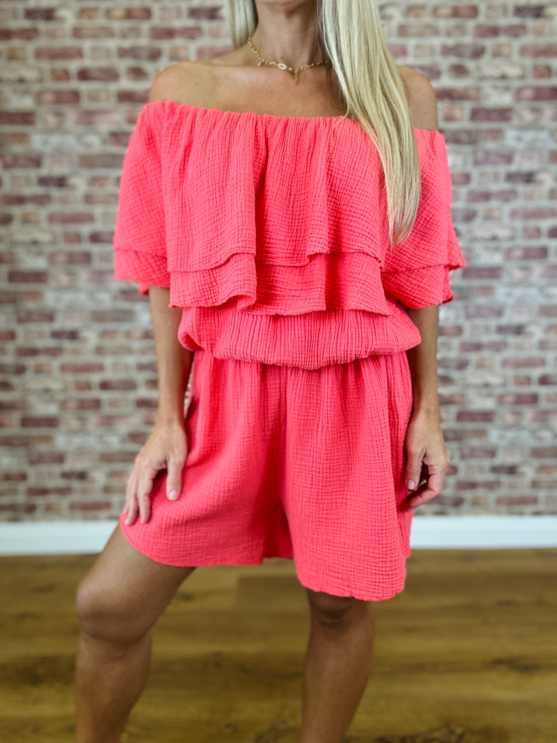 Woman wearing a coral off-shoulder top and shorts set against a brick wall.