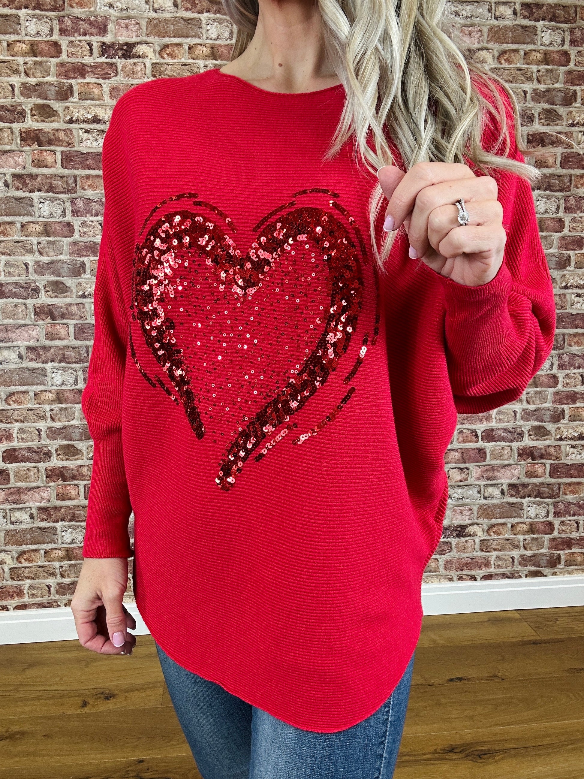Person wearing a red sweater with a sequin heart design in front of a brick wall.