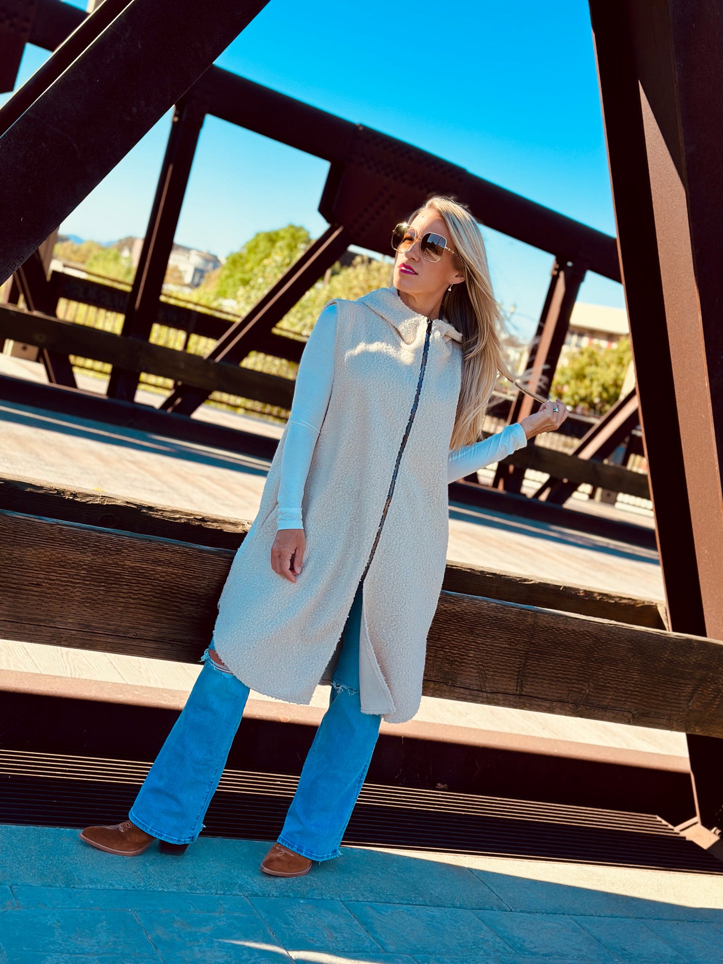 Woman wearing a long beige vest over a light blue top and jeans, standing on a wooden bridge.