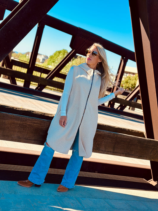 Woman wearing a long beige vest over a light blue top and jeans, standing on a wooden bridge.