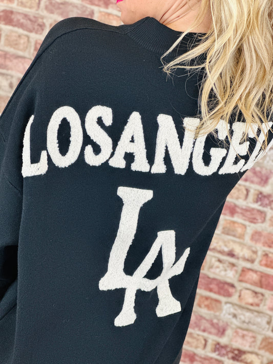 Black sweatshirt with 'Los Angeles' and 'LA' text against a brick wall background