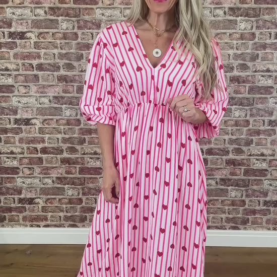 Woman wearing a pink dress with red heart patterns against a brick wall.