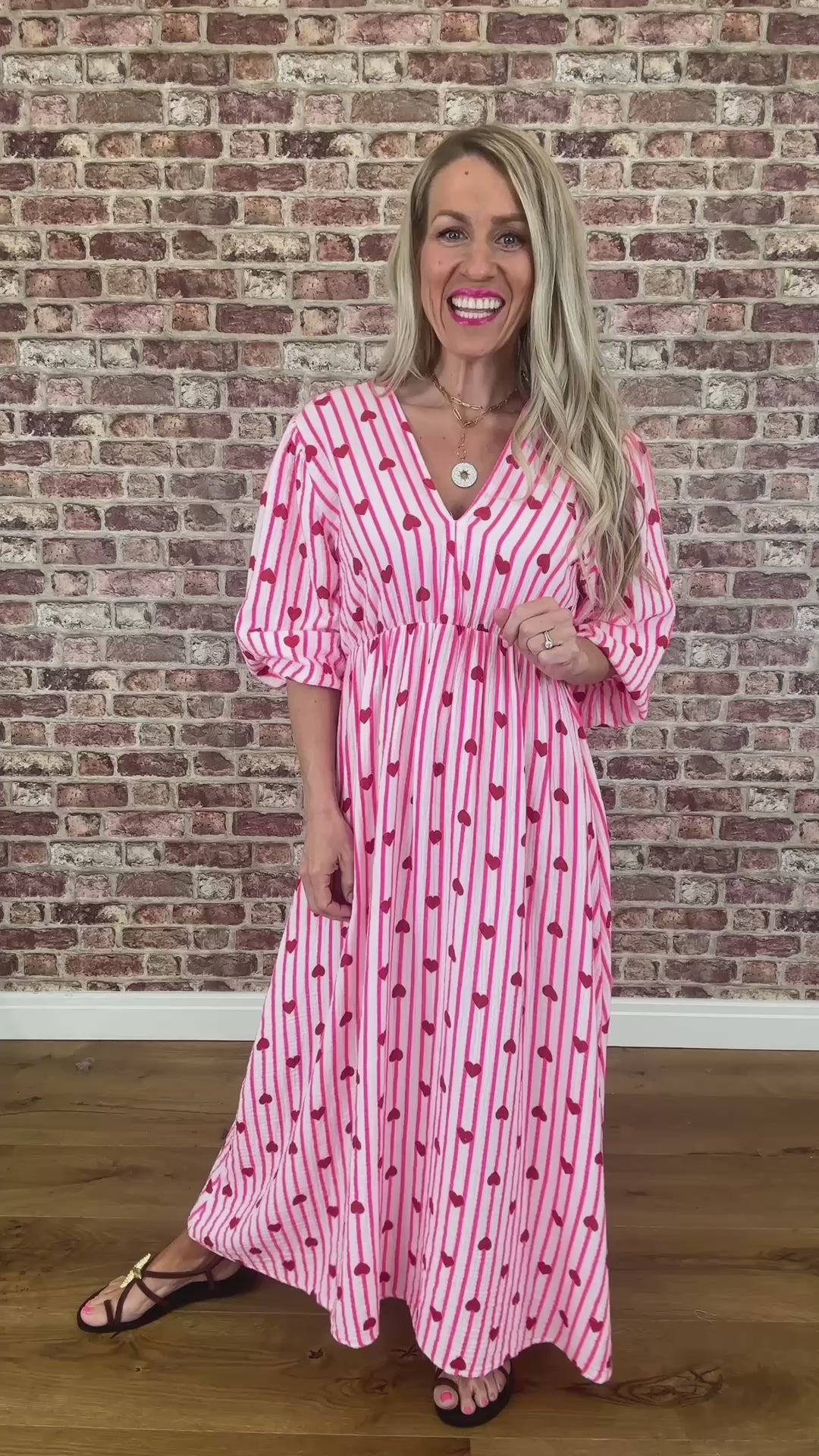Woman wearing a pink dress with red heart patterns against a brick wall.