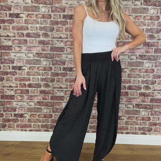 A woman standing against a brick wall backdrop, wearing a white tank top and black wide-leg trousers with side openings, paired with heels.
