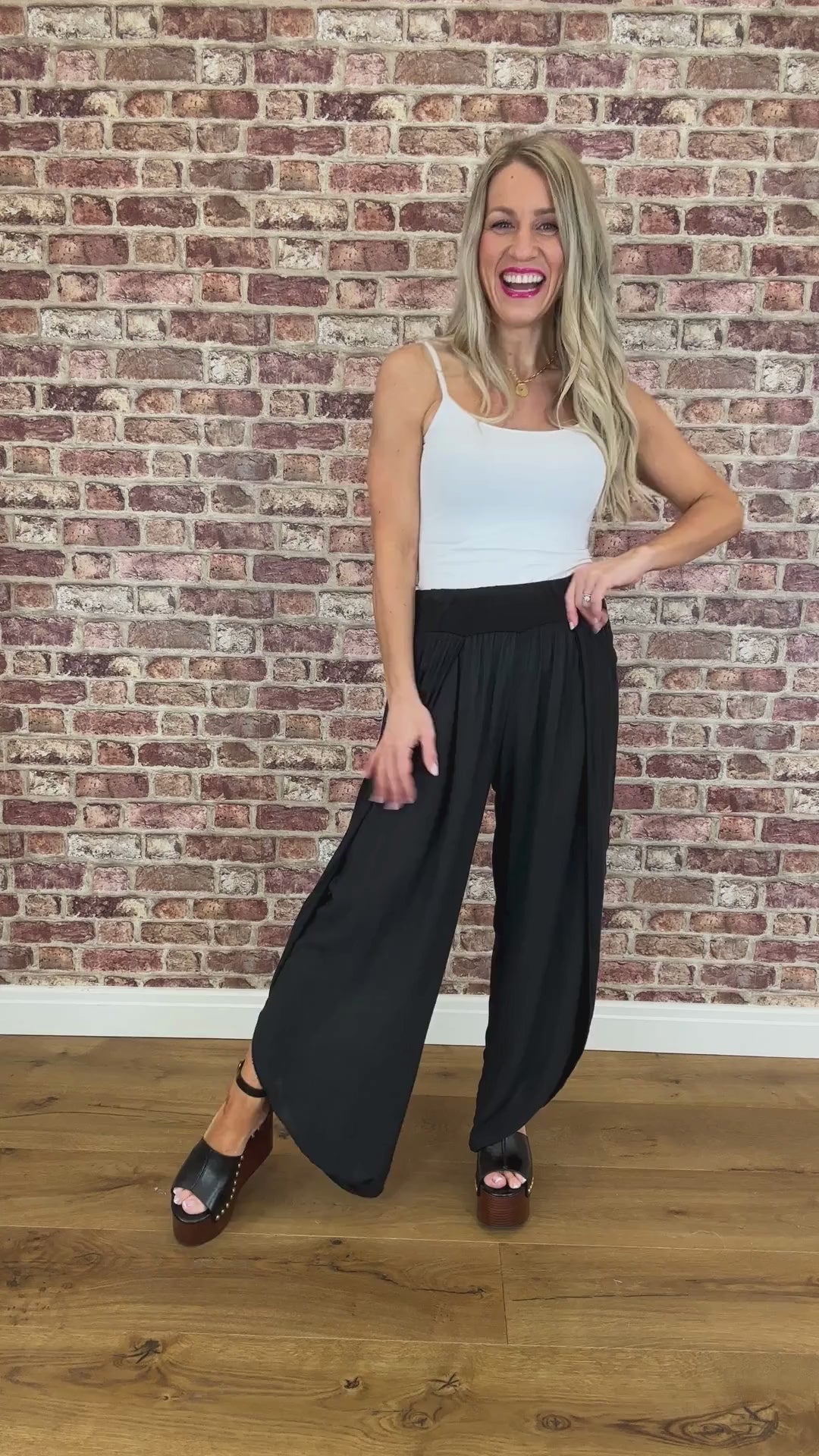 A woman standing against a brick wall backdrop, wearing a white tank top and black wide-leg trousers with side openings, paired with heels.