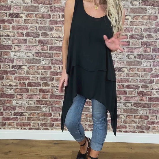 A woman wearing a black sleeveless, V-neck, flowy top with a longer back, paired with blue jeans. She is standing in front of a brick wall.