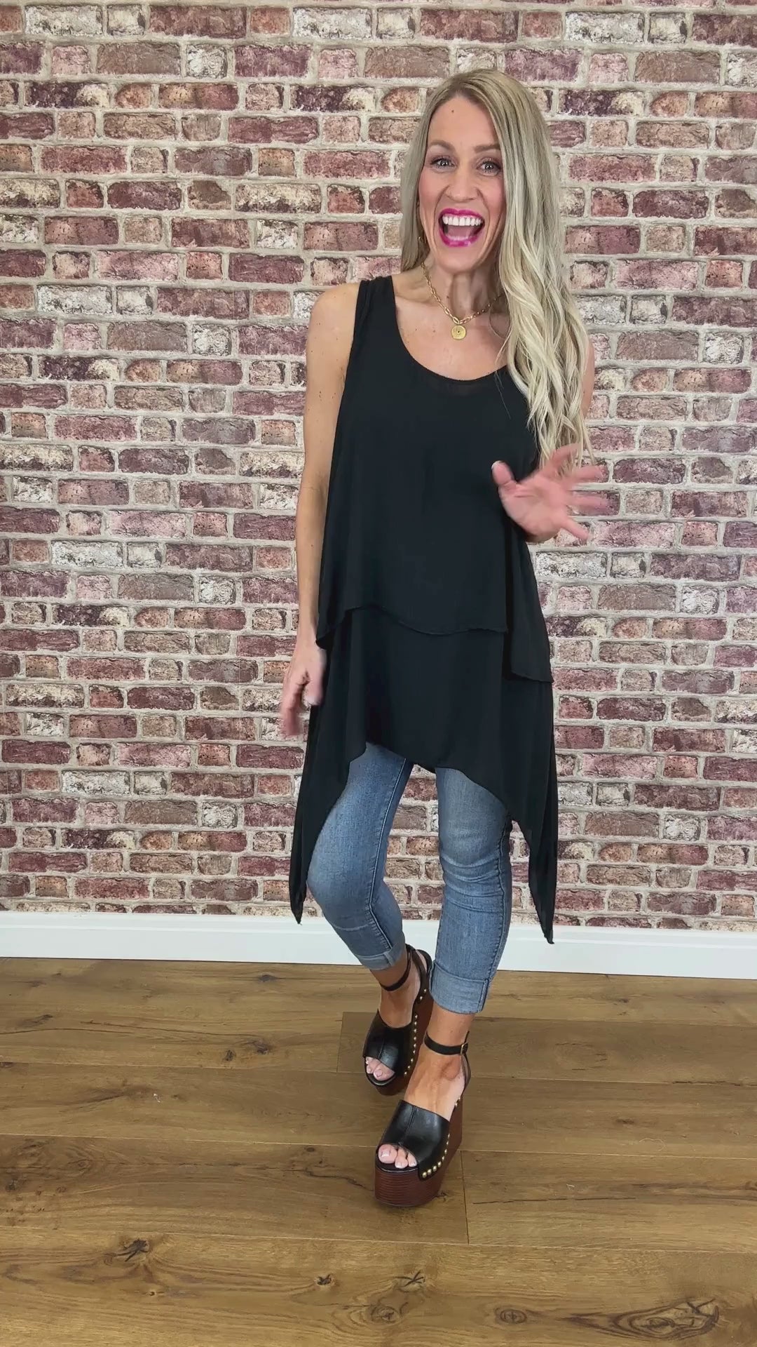A woman wearing a black sleeveless, V-neck, flowy top with a longer back, paired with blue jeans. She is standing in front of a brick wall.