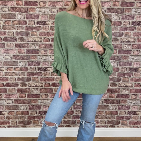 Woman wearing a green sweater and blue jeans standing against a brick wall.