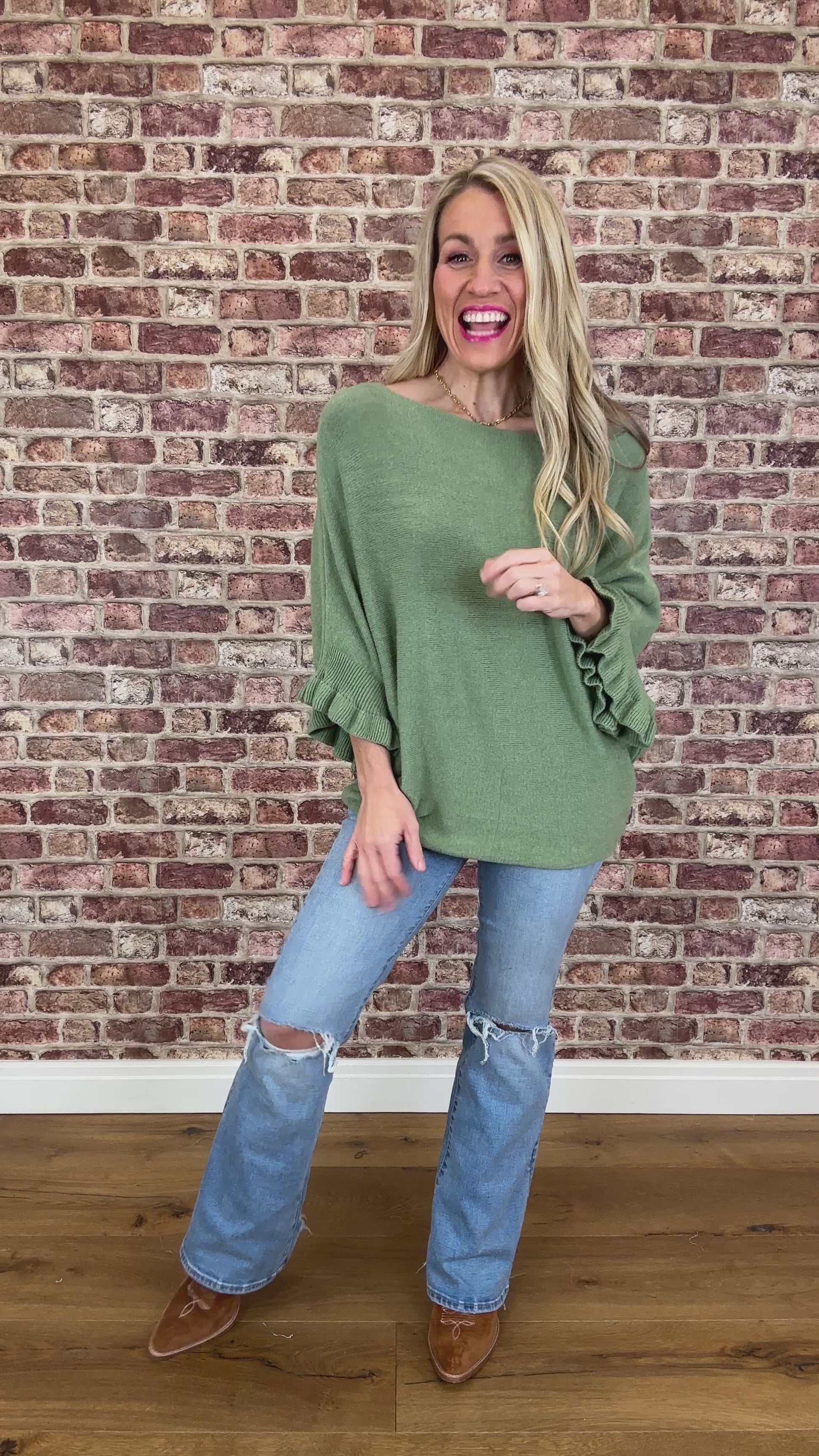 Woman wearing a green sweater and blue jeans standing against a brick wall.