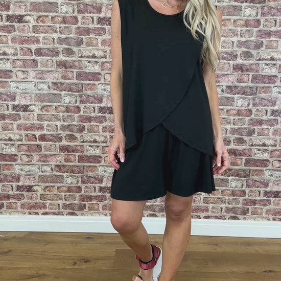 A model wearing a black sleeveless, layered top with wide straps and shorts standing against a brick wall background.