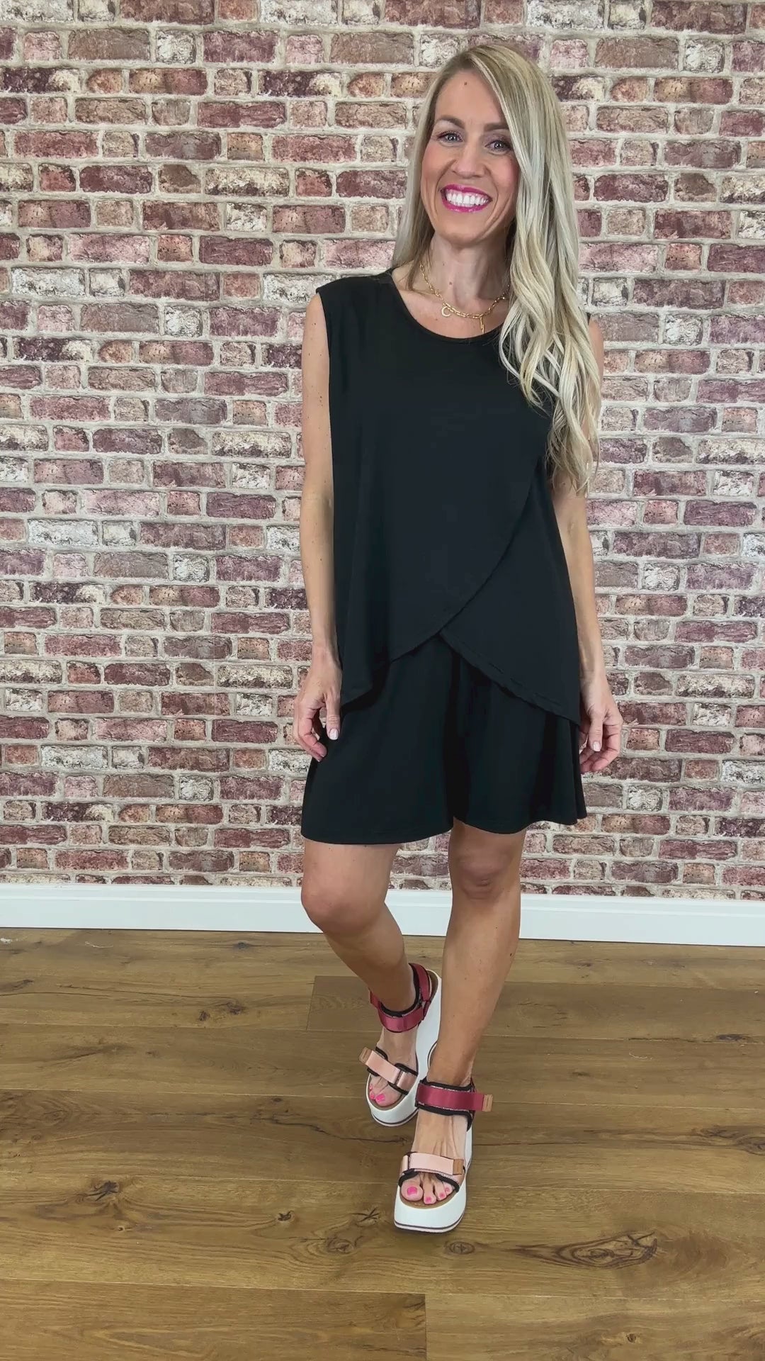 A model wearing a black sleeveless, layered top with wide straps and shorts standing against a brick wall background.