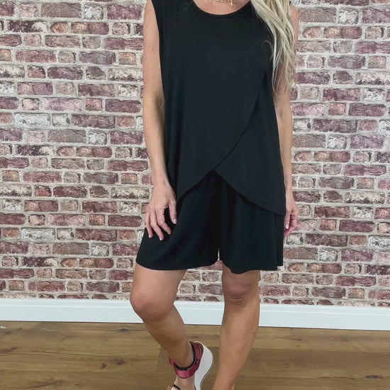 A model wearing black sleeveless shorts with a flowing asymmetrical design, standing against a brick wall background.