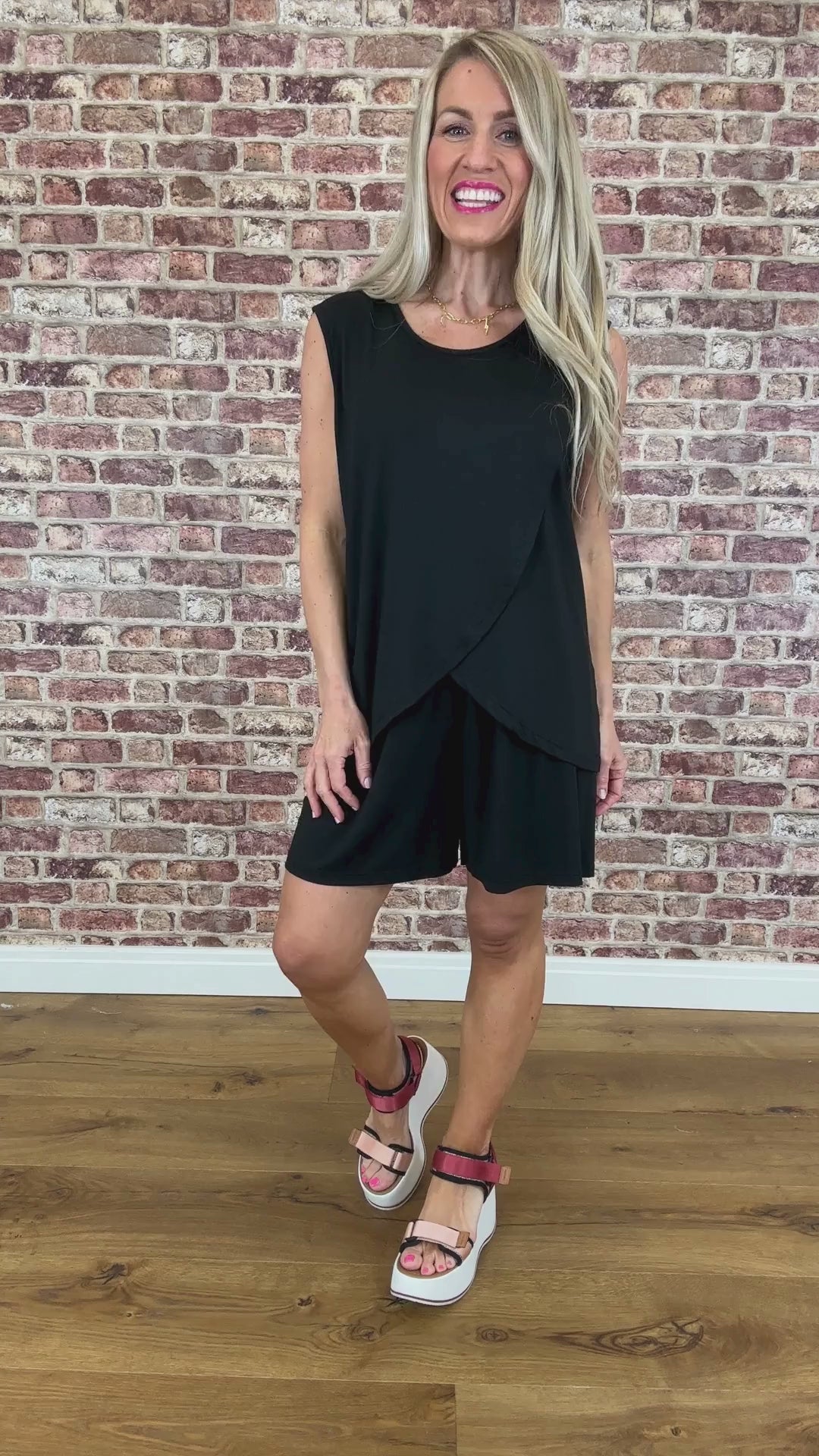 A model wearing black sleeveless shorts with a flowing asymmetrical design, standing against a brick wall background.